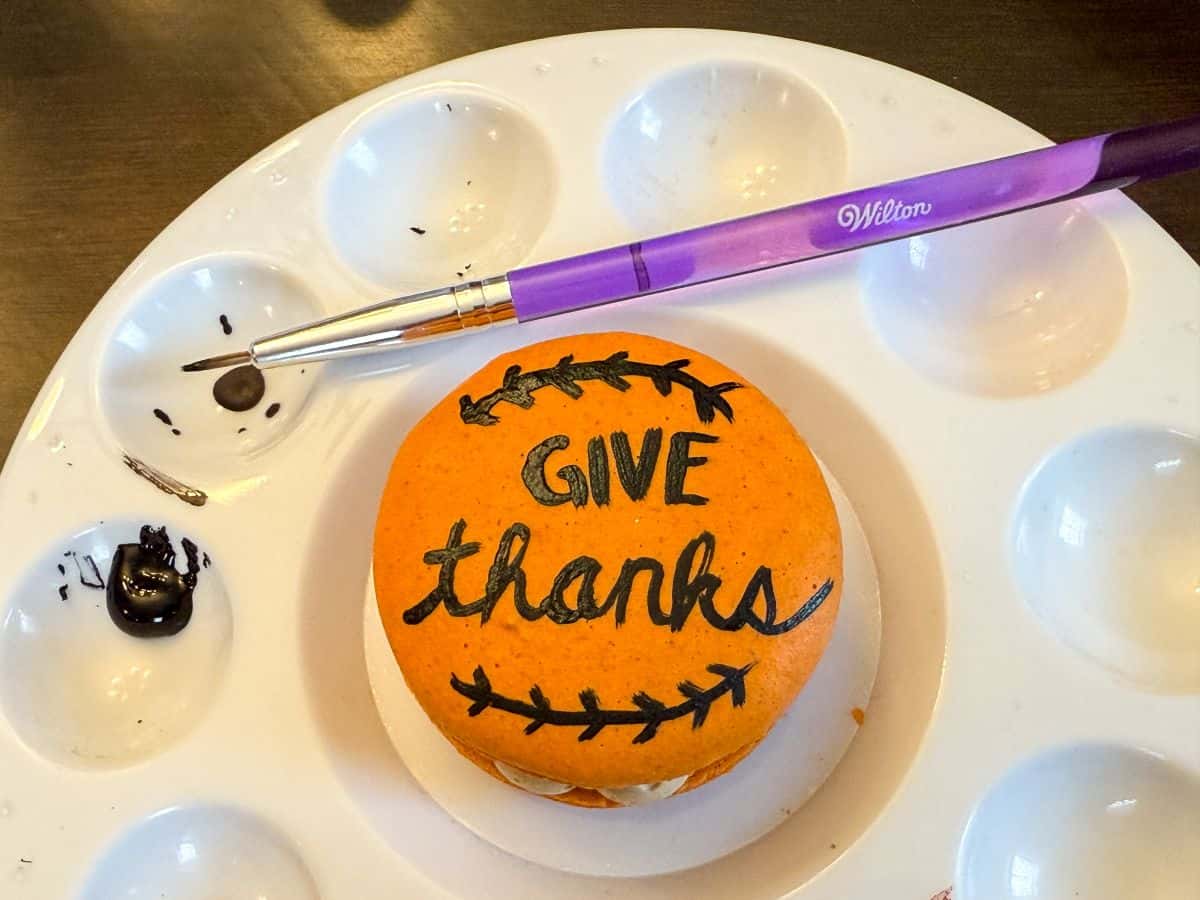 an orange macaron in the center of a paint palette with the words "Give thanks" painted on using black food coloring gel
