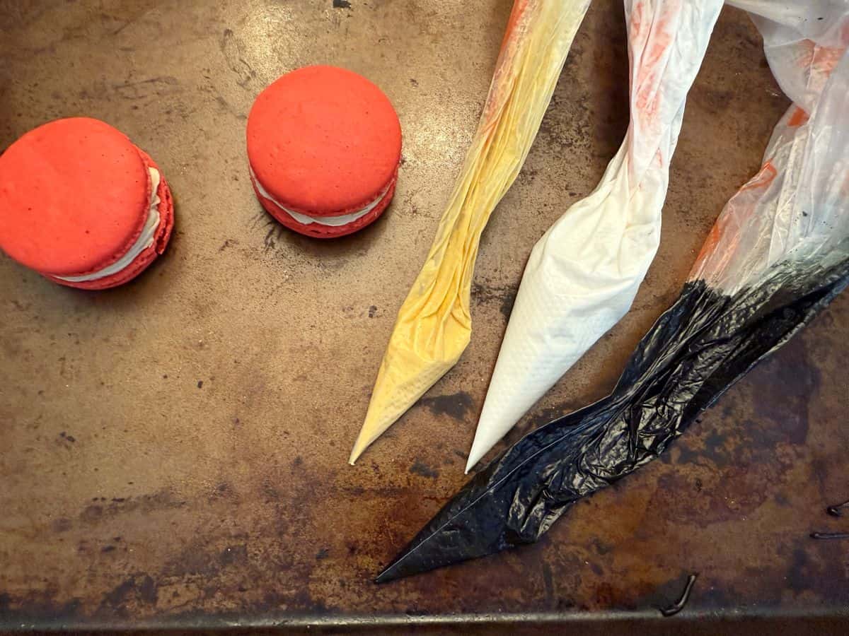 two red macarons next to piping bags filled with gold, white and black royal icing