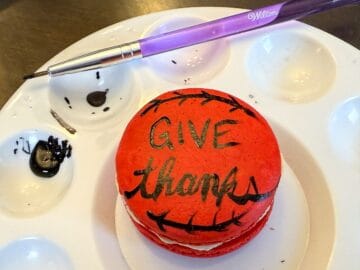 a red macaron in the center of a paint palette with the words "Give thanks" painted on using black food coloring gel