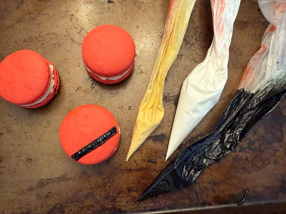 red macarons, one with a black royal icing Santa belt, next to piping bags filled with gold, white and black royal icing