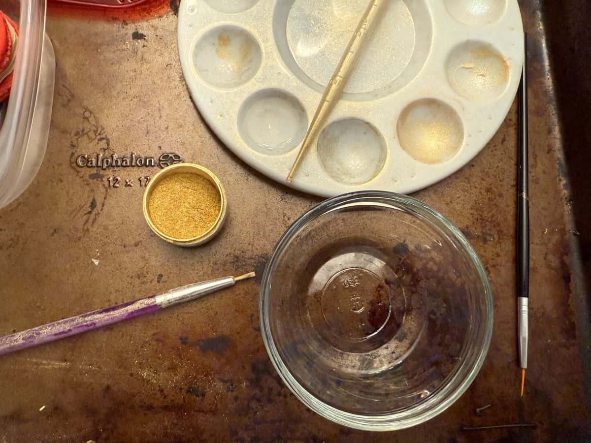 a paint palette next to a container of edible gold luster dust and a bowl of Everclear with a small food grade paintbrush and disposable transfer pipette
