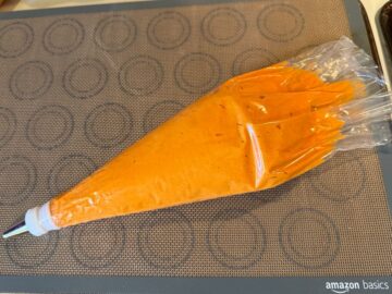 orange nut free macaron batter in a disposable piping bag fitted with a medium round tip on top of a macaron mat