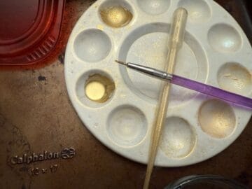 a paint palette with a well filled with gold luster dust suspension next to a food grade paintbrush and disposable transfer pipette