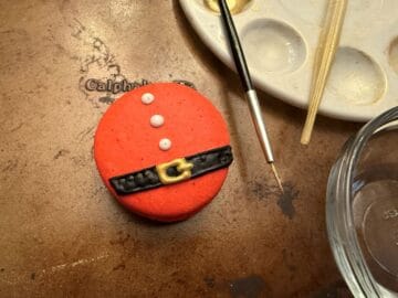 a red macaron with royal icing Santa belt next to a paint palette, small paintbrush and disposable transfer pipette