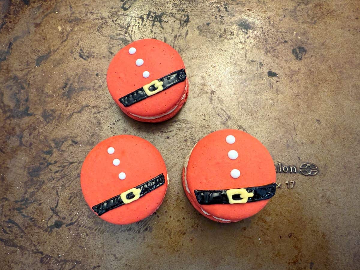 three Santa Macarons with black belts, a gold buckle, and three white buttons before being painted with gold luster dust