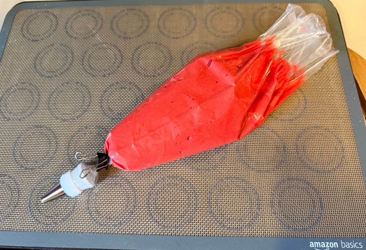 red macaron batter in a disposable piping bag fitted with a medium round tip on top of a macaron mat