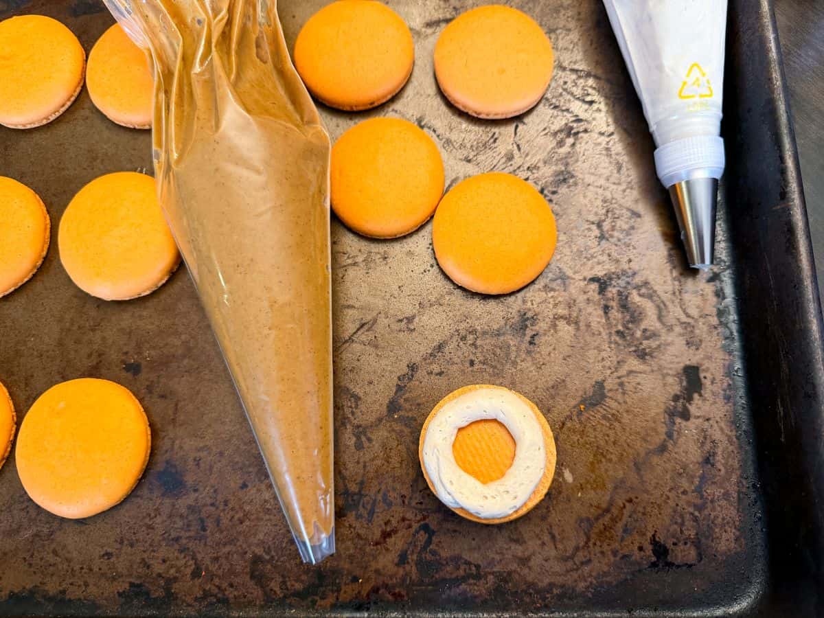 orange macaron shells on a tray next to a piping bag filled with pumpkin filling with one shell having a piped coffee buttercream border