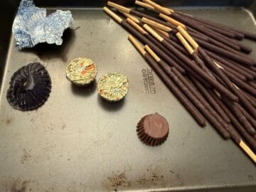 an unwrapped Reese's miniature peanut butter cup next to pocky sticks on a baking sheet