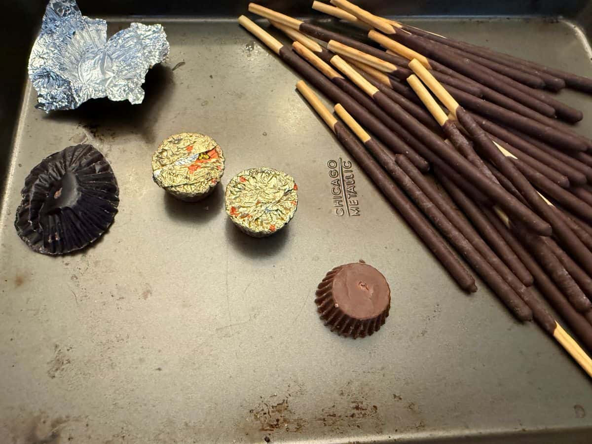 an unwrapped Reese's miniature peanut butter cup next to pocky sticks on a baking sheet