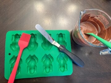 green chocolate frog silicone mold with a silicone pastry brush and small offset spatula next to a glass measuring cup filled with melted chocolate