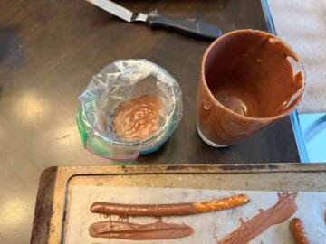 a sandwich bag filled with melted chocolate next to a glass of melted chocolate and pretzel rods on a waxed paper lined cookie sheet
