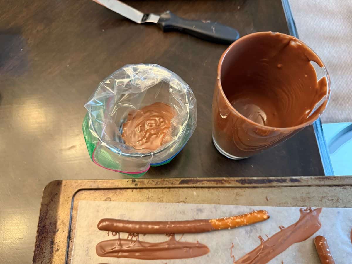 a sandwich bag filled with melted chocolate next to a glass of melted chocolate and pretzel rods on a waxed paper lined cookie sheet