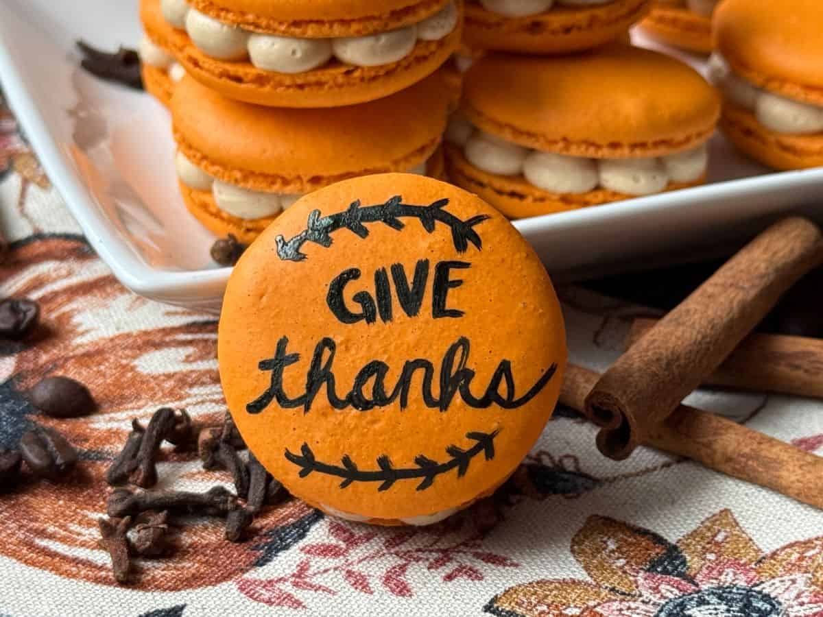 an orange macaron that says "Give thanks" in front of stacked macarons surrounded by cinnamon sticks and cloves