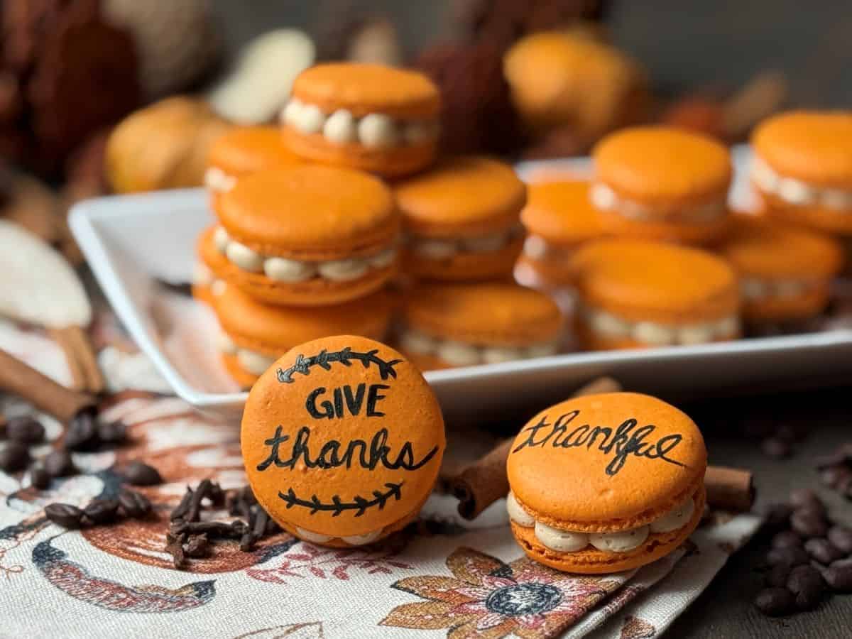Painted Thanksgiving Macarons stacked on a plate and in front of the plate with a fall towel and cinnamon sticks and cloves sprinkled around