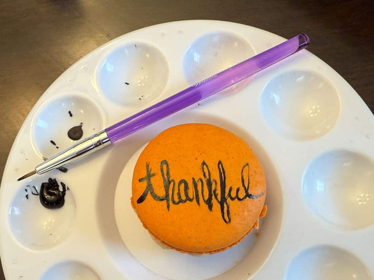 an orange macaron in the center of a paint palette with the word "thankful" painted on with black food coloring gel