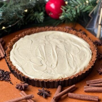 an eggnog tart with gingersnap crust on a wood surface with holiday foliage, cinnamon sticks, and star anise around it