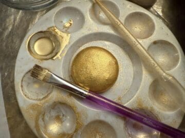 a gold painted macaron shell in the center of a paint palette with edible gold luster dust suspension in a well