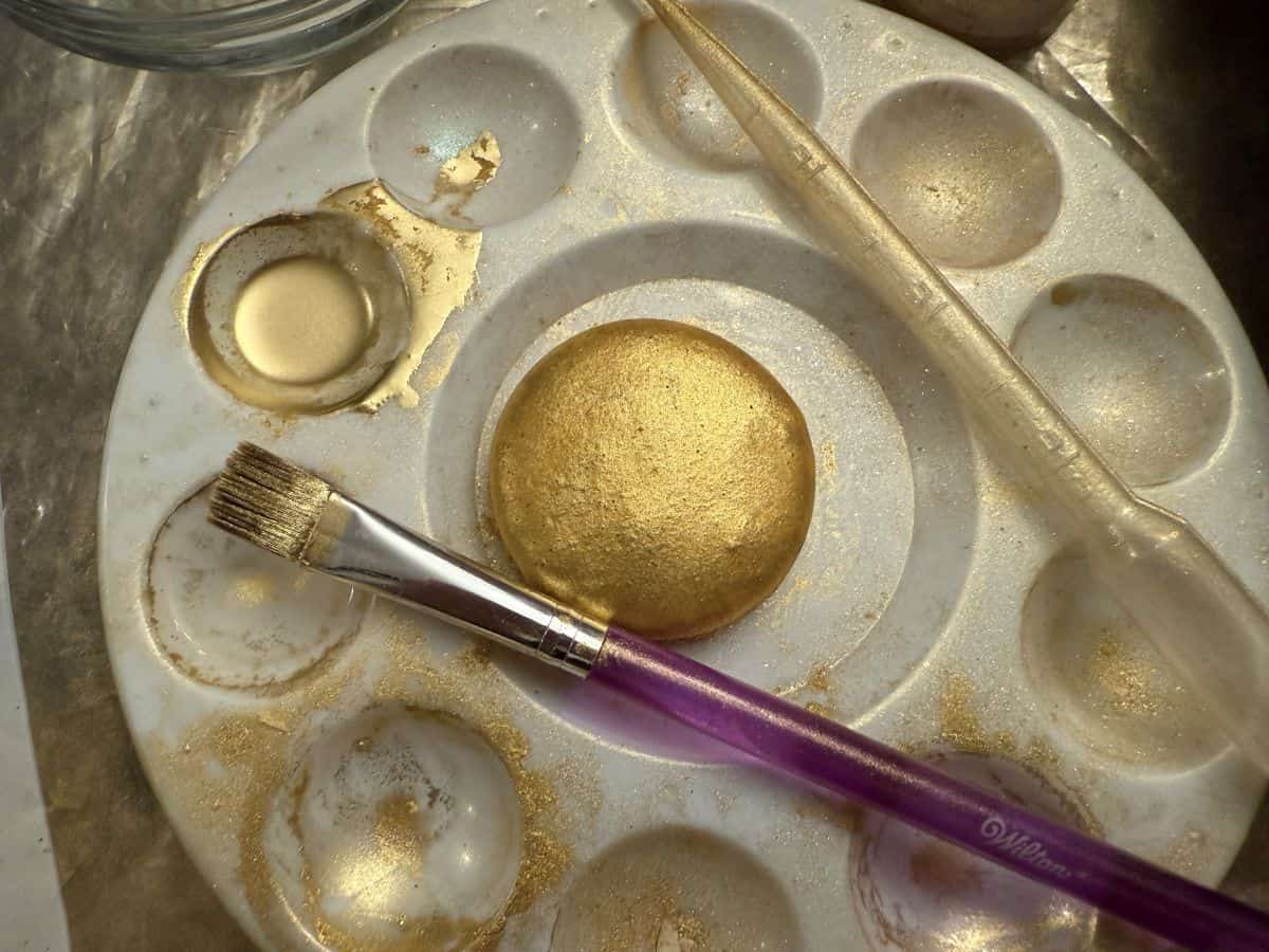 a gold painted macaron shell in the center of a paint palette with edible gold luster dust suspension in a well
