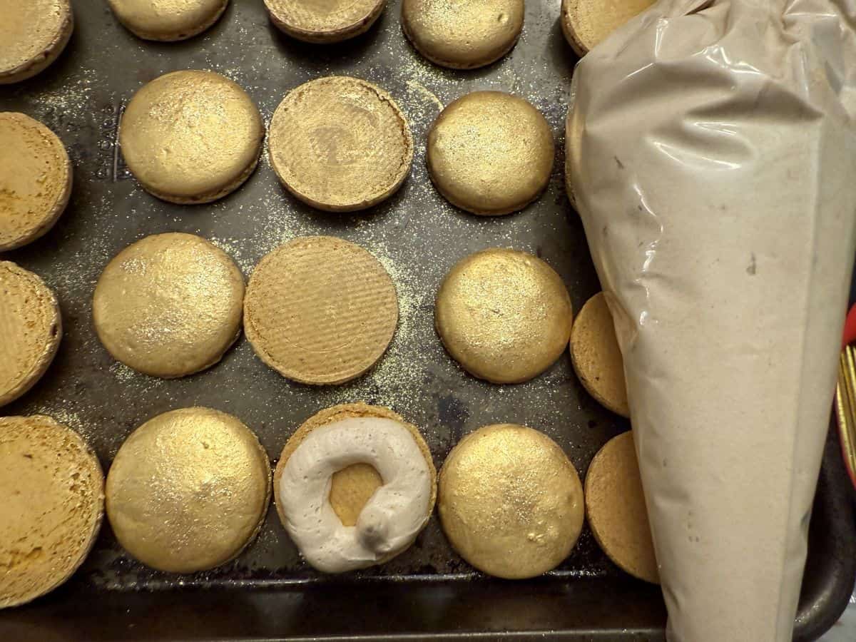 gold macaron shells on a baking sheet next to a piping bag filled with butterscotch buttercream