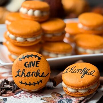 Painted Thanksgiving Macarons stacked on a plate and in front of the plate with a fall towel and cinnamon sticks and cloves sprinkled around
