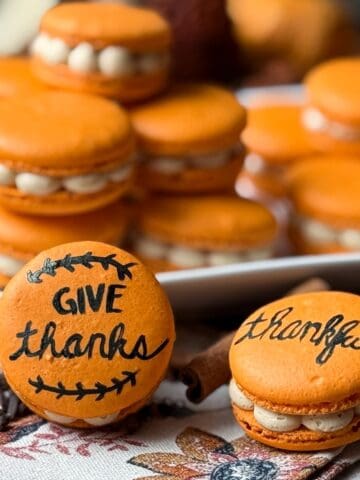 Painted Thanksgiving Macarons stacked on a plate and in front of the plate with a fall towel and cinnamon sticks and cloves sprinkled around