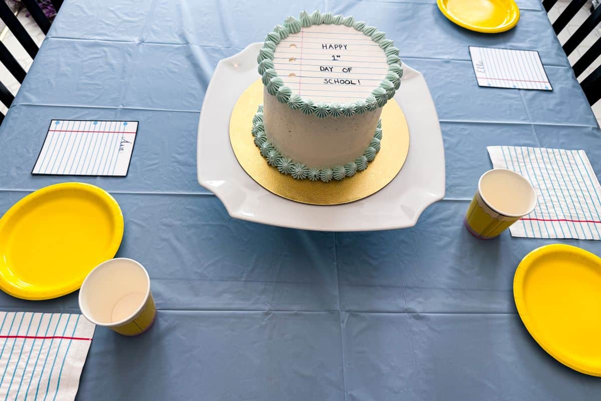 a First Day of School Cake in the center of a blue tablecloth covered table with yellow plates