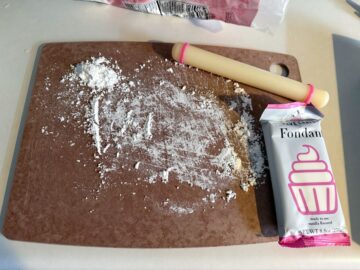 a cutting board dusted with powdered sugar with a small fondant rolling pin and package of white fondant