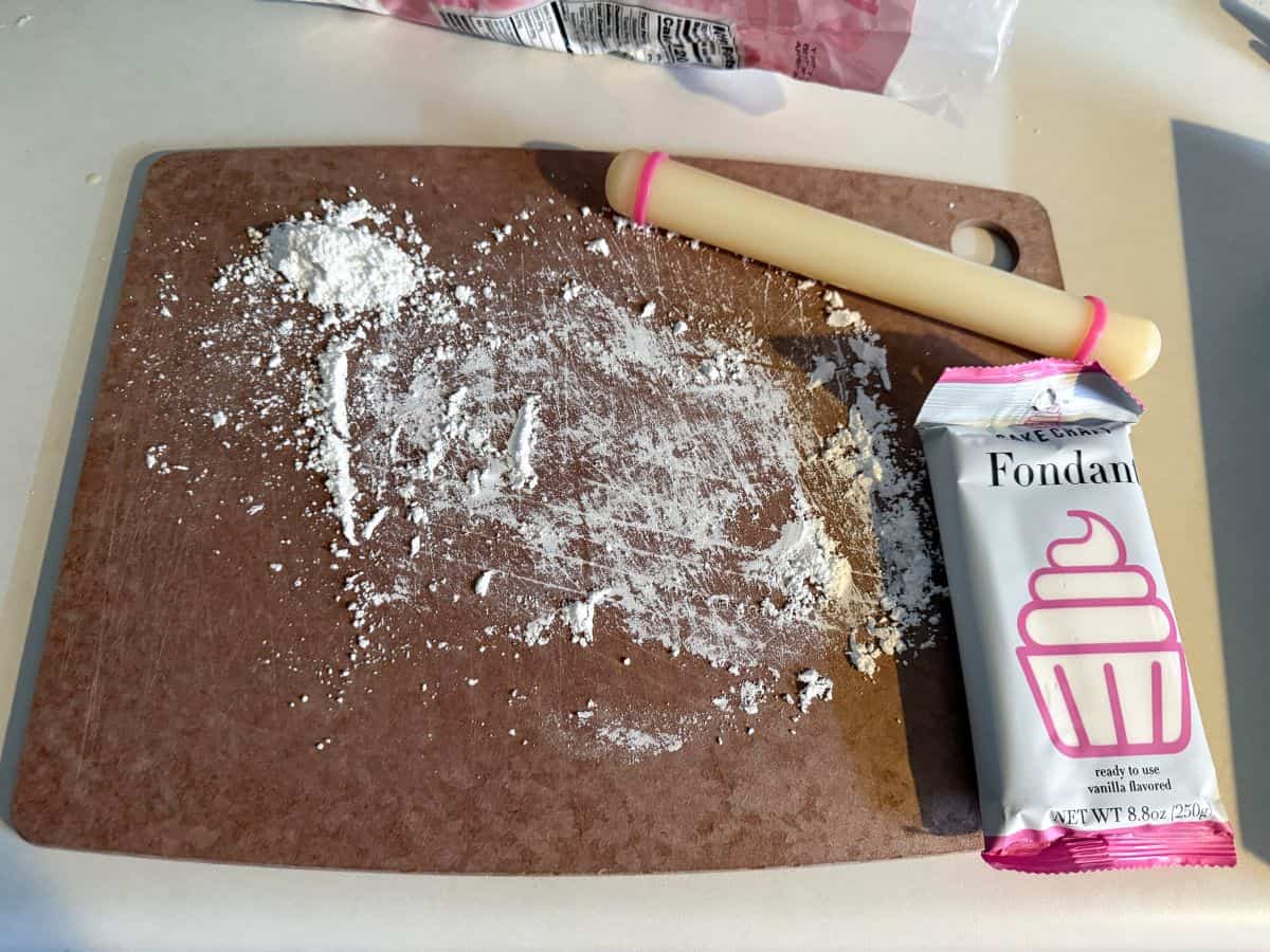 a cutting board dusted with powdered sugar with a small fondant rolling pin and package of white fondant