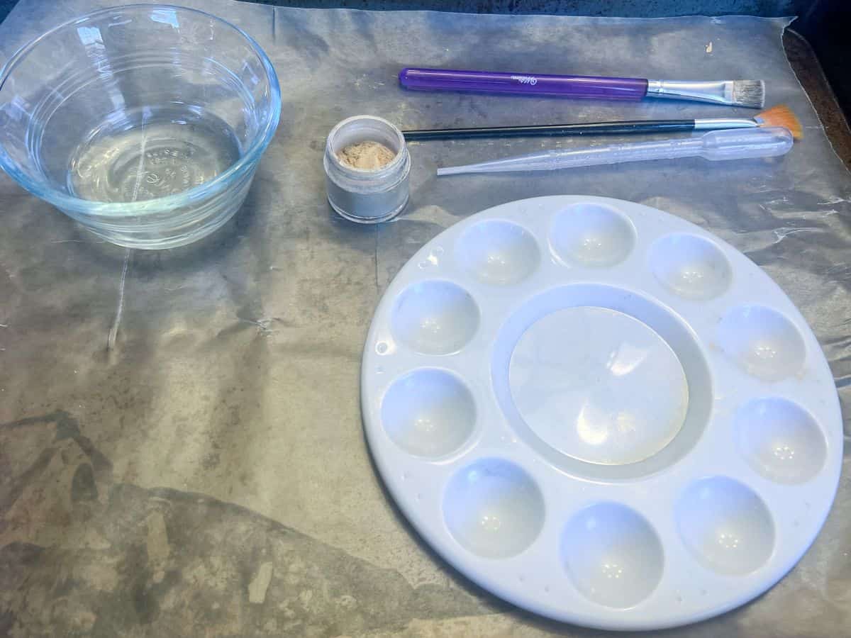 a paint palette with paintbrushes, gold luster dust, and a bowl filled with clear alcohol