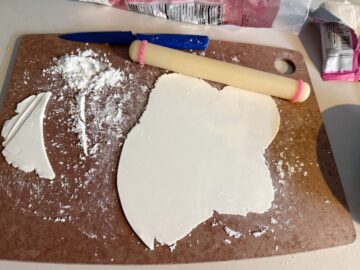 a piece of white fondant rolled into a thin layer with a small fondant rolling pin on a cutting board