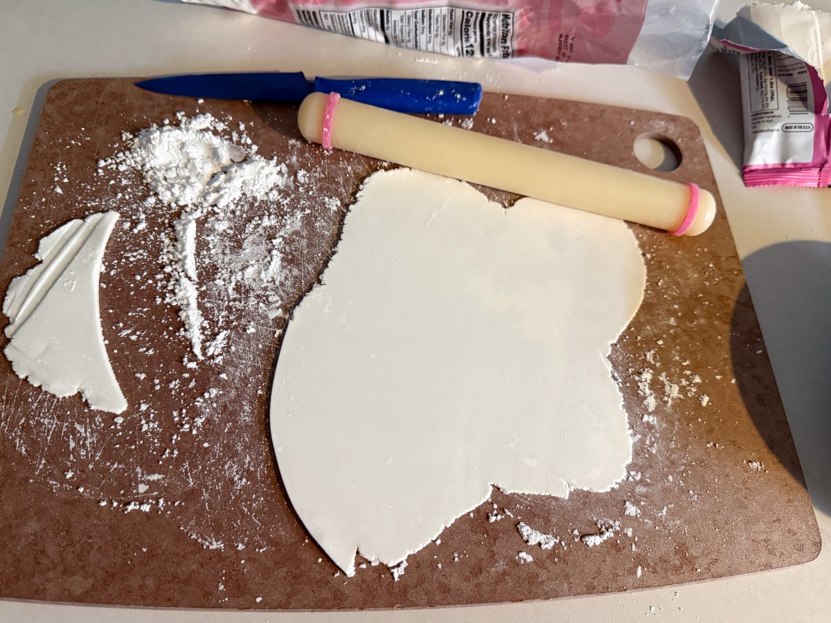 a piece of white fondant rolled into a thin layer with a small fondant rolling pin on a cutting board