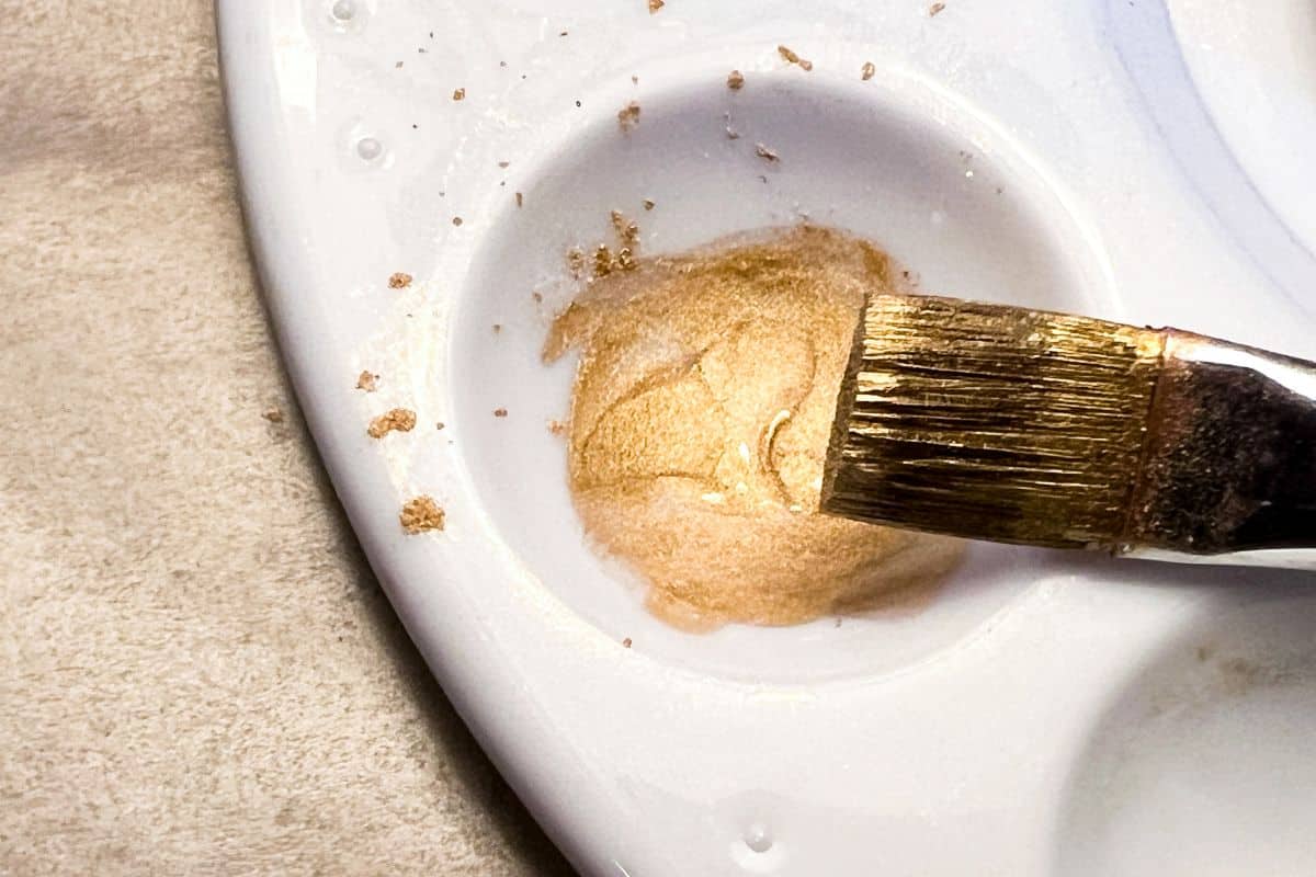 gold luster dust suspension in the well of a paint palette with a gold covered paintbrush held up