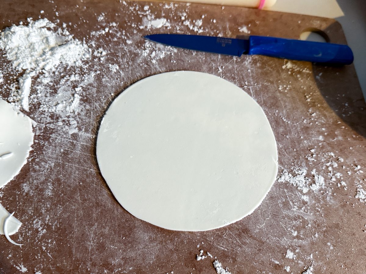 a white rolled fondant circle on a powdered sugar dusted cutting board with a paring knife