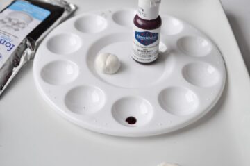 white fondant shaped into a ladybug next to a bottle of red food coloring gel on a food dedicated paint palette