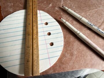 a wood ruler on top of a round piece of white rolled fondant that has blue parallel lines and a perpendicular pink line drawn with food coloring markers