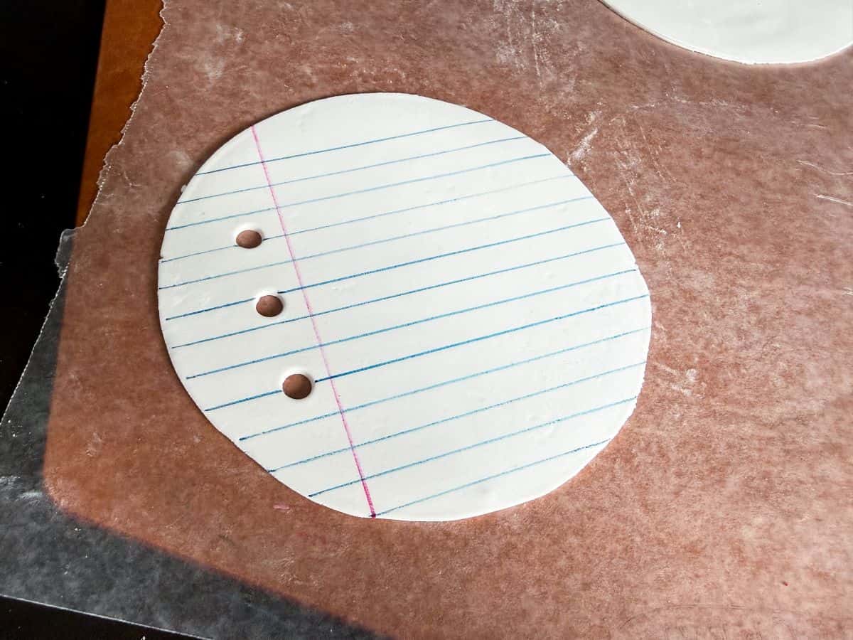 a round piece of white rolled fondant that has blue parallel lines and a perpendicular pink line drawn with food coloring markers to look like a piece of loose leaf notebook paper