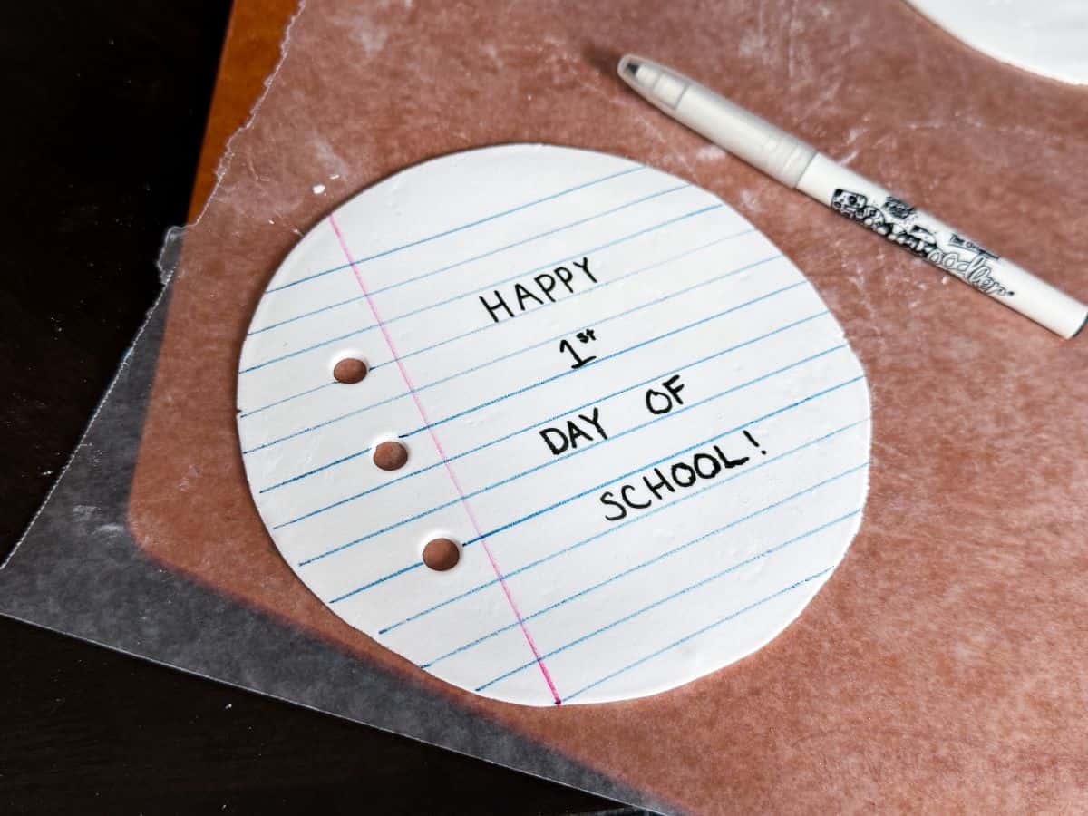 a round piece of white rolled fondant that has blue parallel lines and a perpendicular pink line drawn with food coloring markers to look like a piece of loose leaf notebook paper with black writing that says "Happy 1st Day of School!"