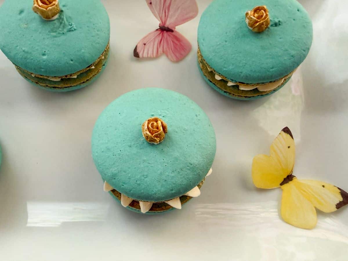 gold mini buttercream roses on top of macarons on a white plate surrounded by edible butterflies