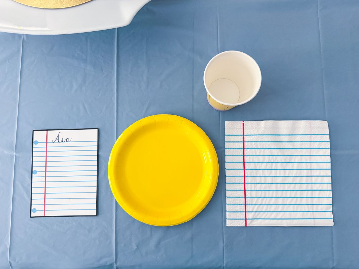 a First Day of School place setting that has a yellow paper plate, a notebook paper napkin, a yellow pencil paper cup and a notebook paper name card