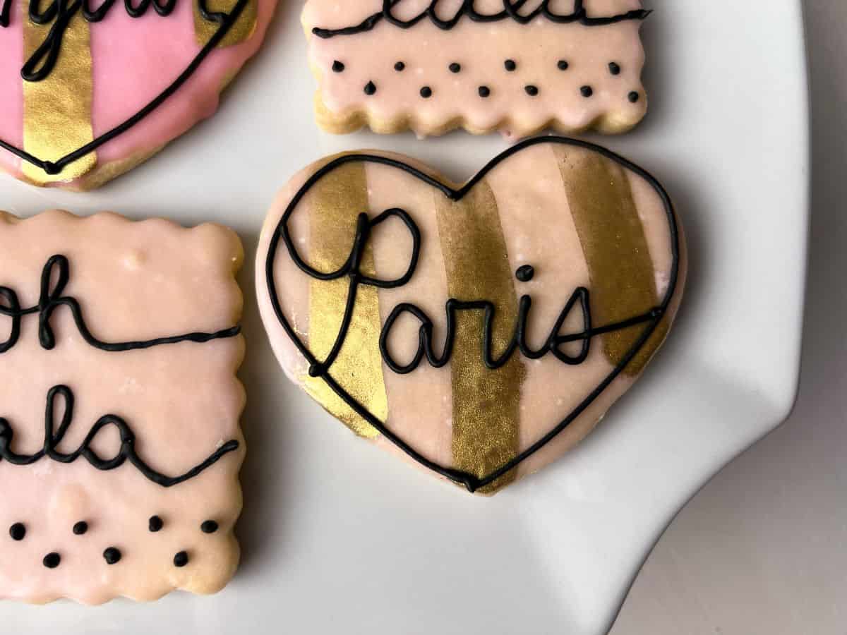 a heart shaped cookie that is light pink with gold stripes painted on and black royal icing writing that says "Paris"